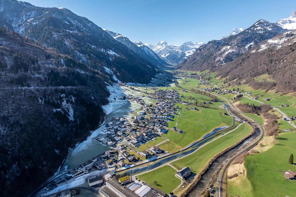 Luftaufnahme eines alpinen Tals mit Fluss, Ortschaft und Bergen