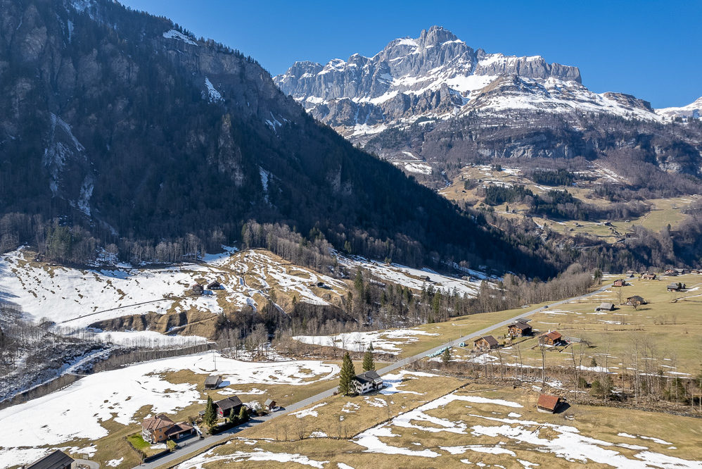 Alpenlandschaft mit Tal, vereinzelt stehenden Häusern und schneebedeckten Bergen