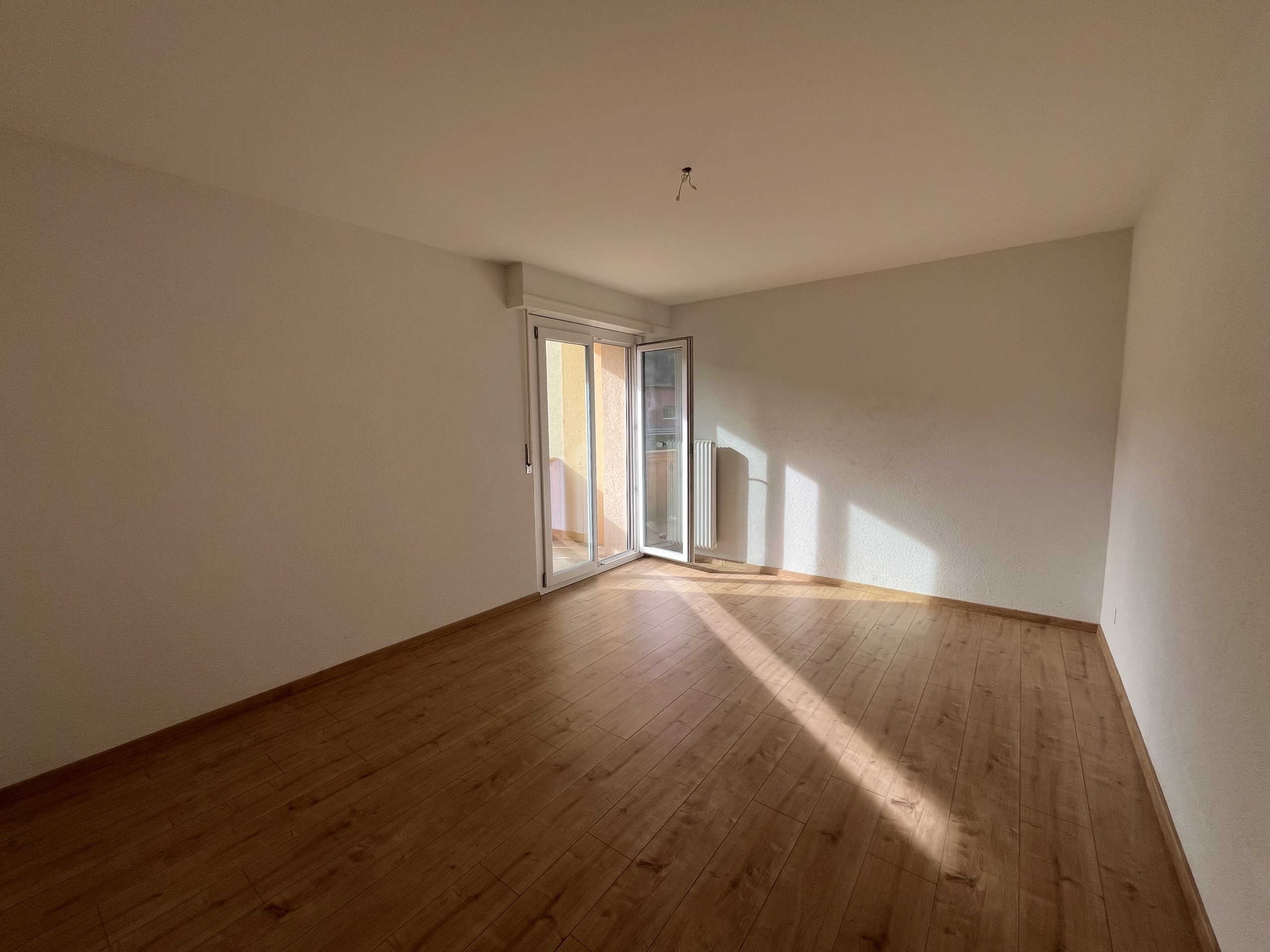 Leerer, sonnendurchfluteter Raum mit Holzboden und Balkonzugang