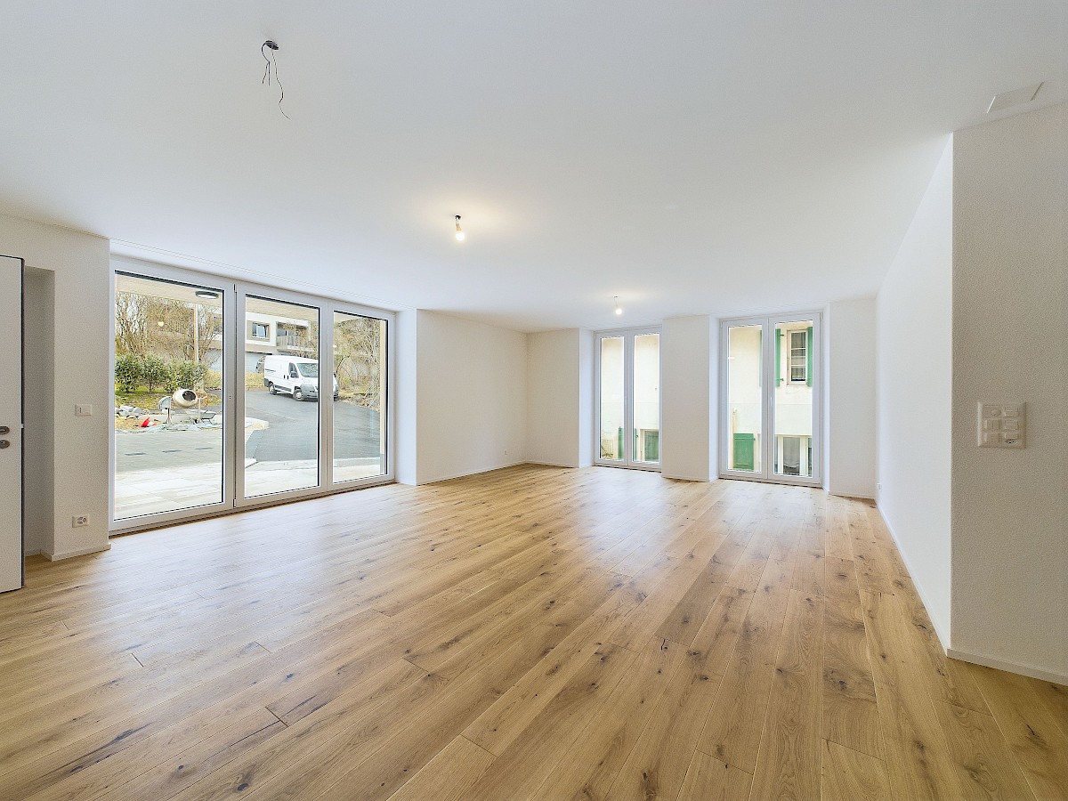 Großer leerer Wohnraum mit Holzoptik-Boden und bodentiefen Fenstern
