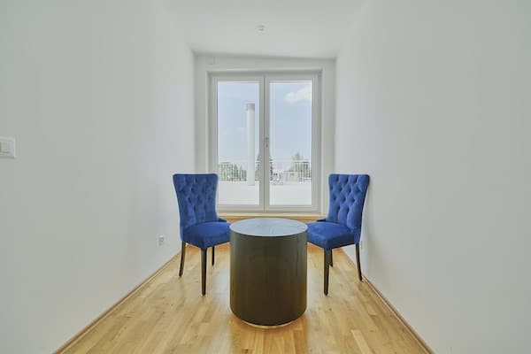 Heller, leerer Raum mit zwei blauen Stühlen vor großem Fenster