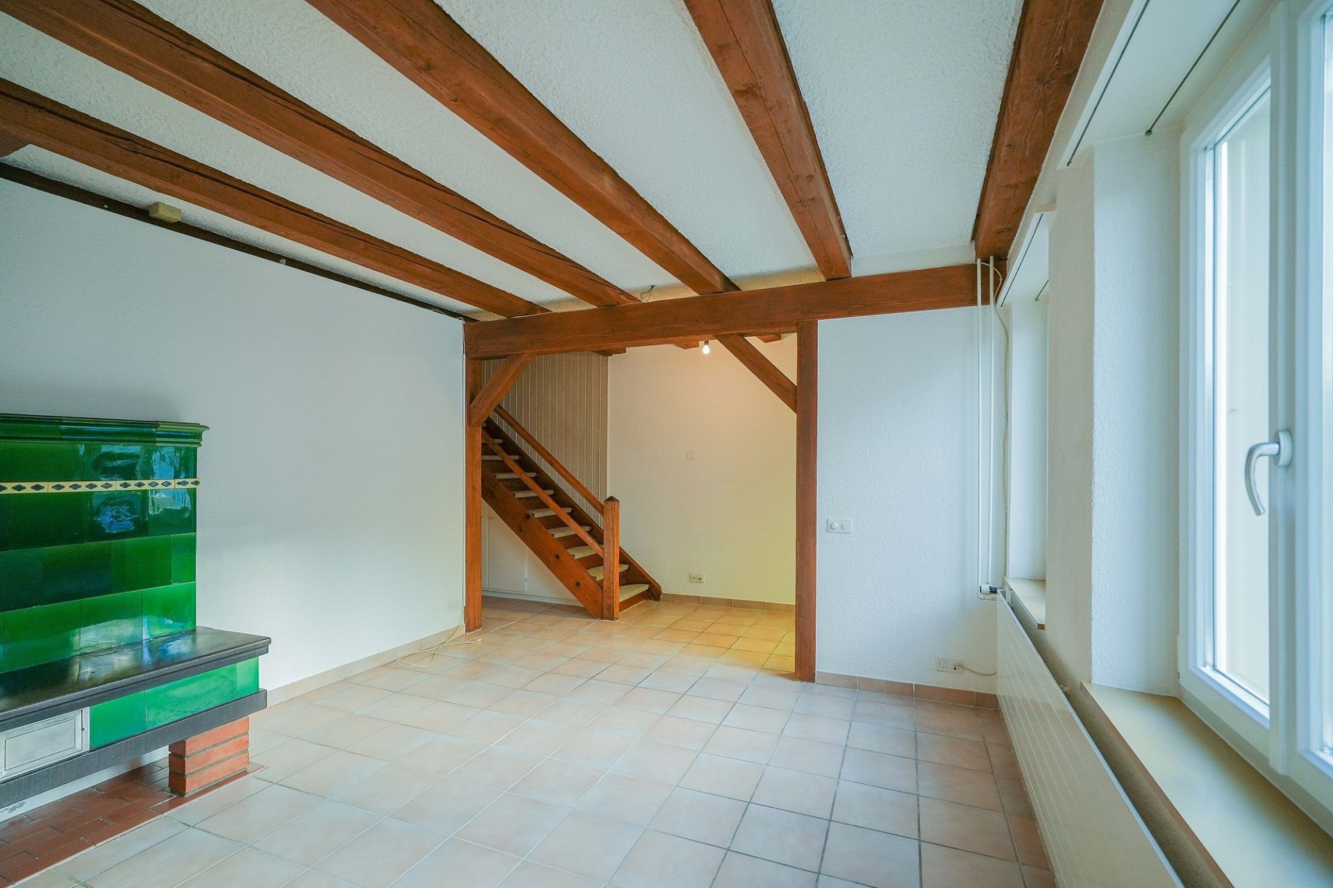 Großer leerer Wohnraum mit Kachelofen und sichtbaren Holzbalken