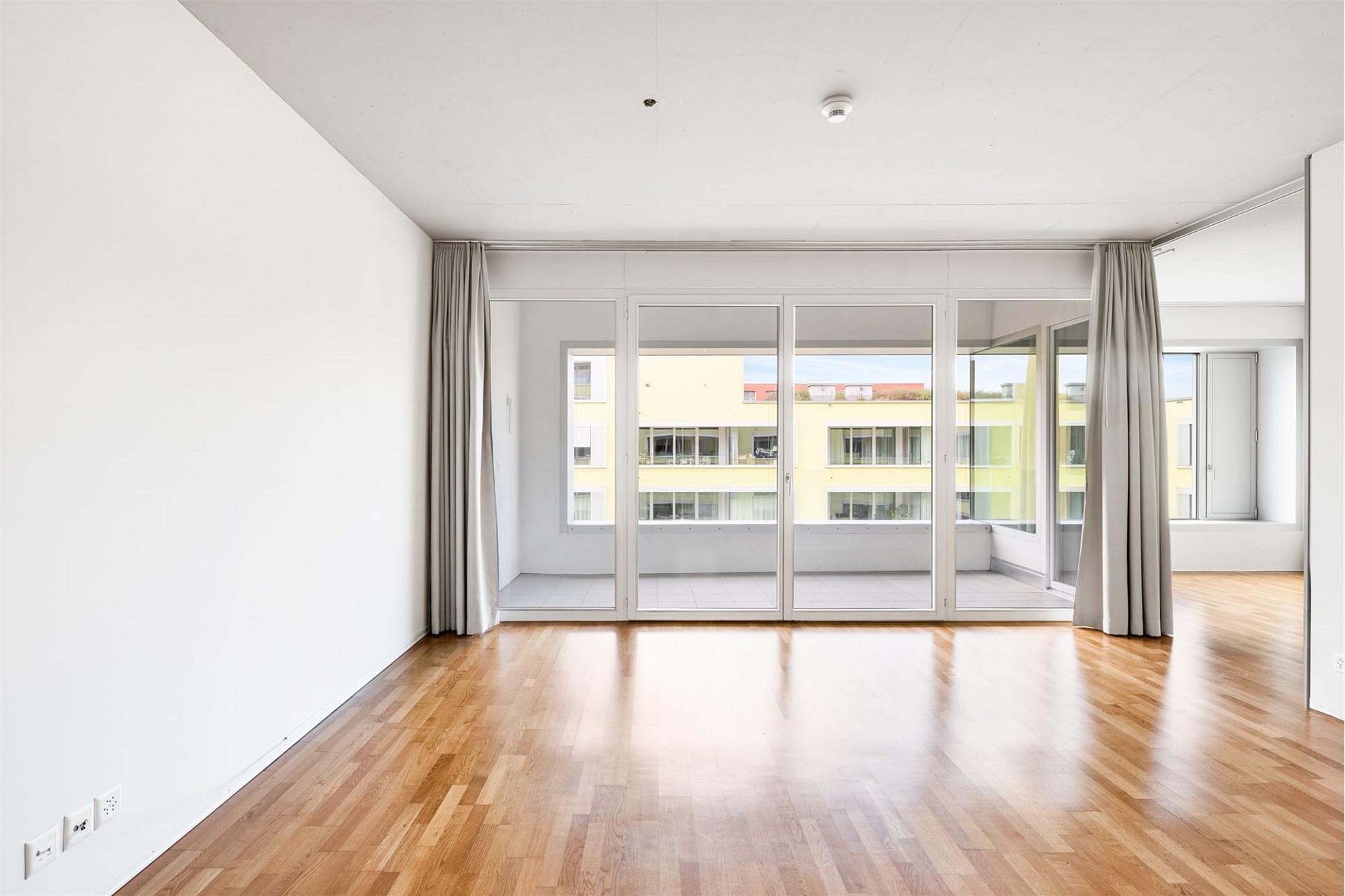 Helles, leeres Wohnzimmer mit Balkon und Blick ins gegenüberliegende Gebäude