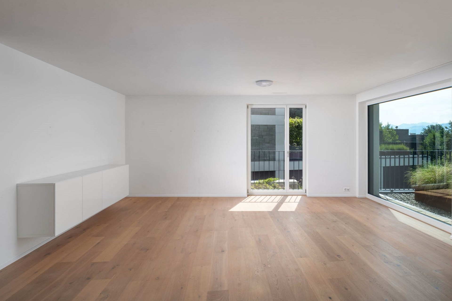 Leerer, modern wirkender Wohnraum mit Holzparkett und großen Fensterflächen