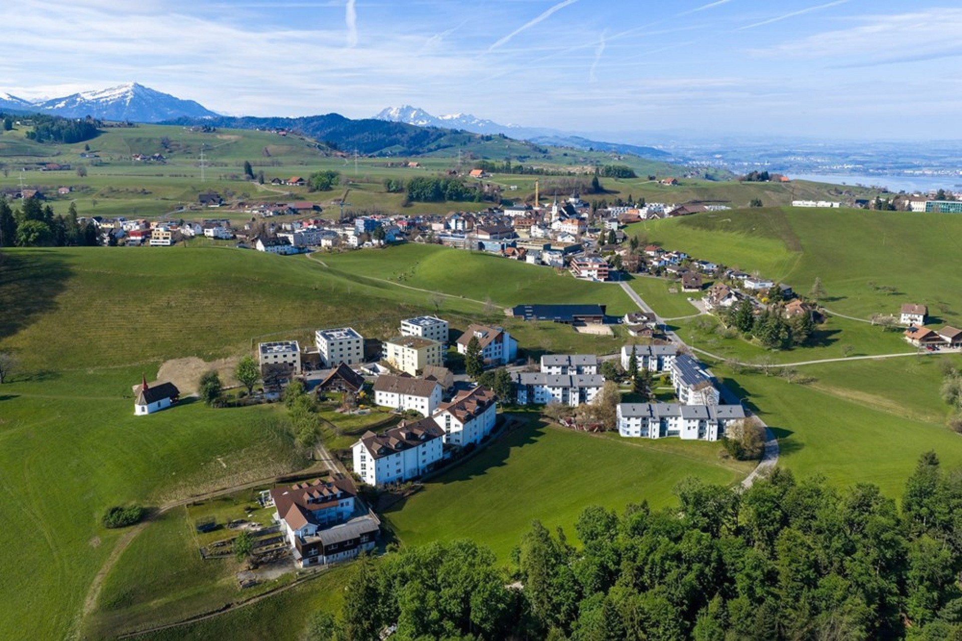 Luftaufnahme einer grünen Hügellandschaft mit Siedlung und Bergpanorama