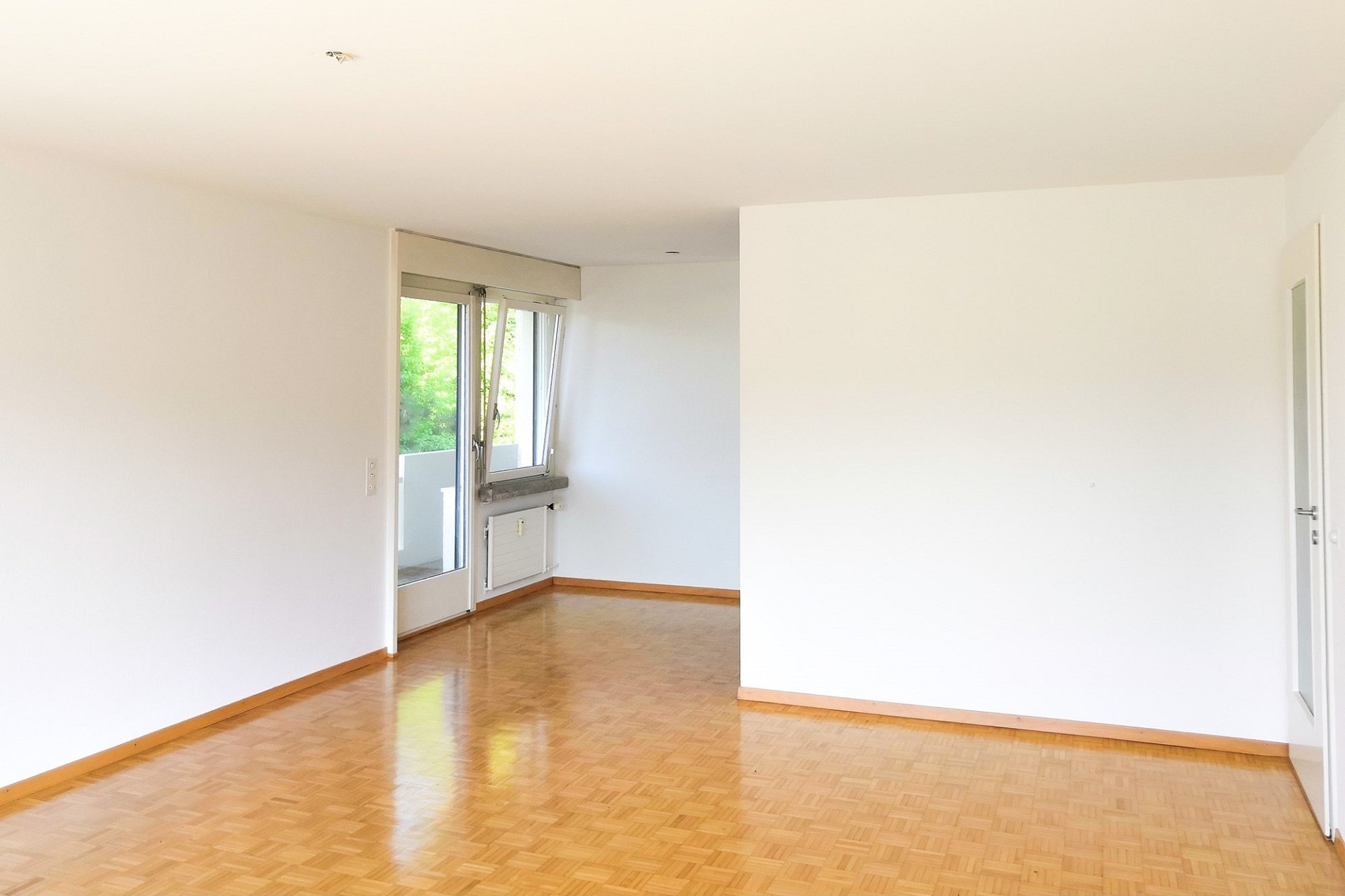 Heller leerer Wohnraum mit Parkettboden und Balkonzugang