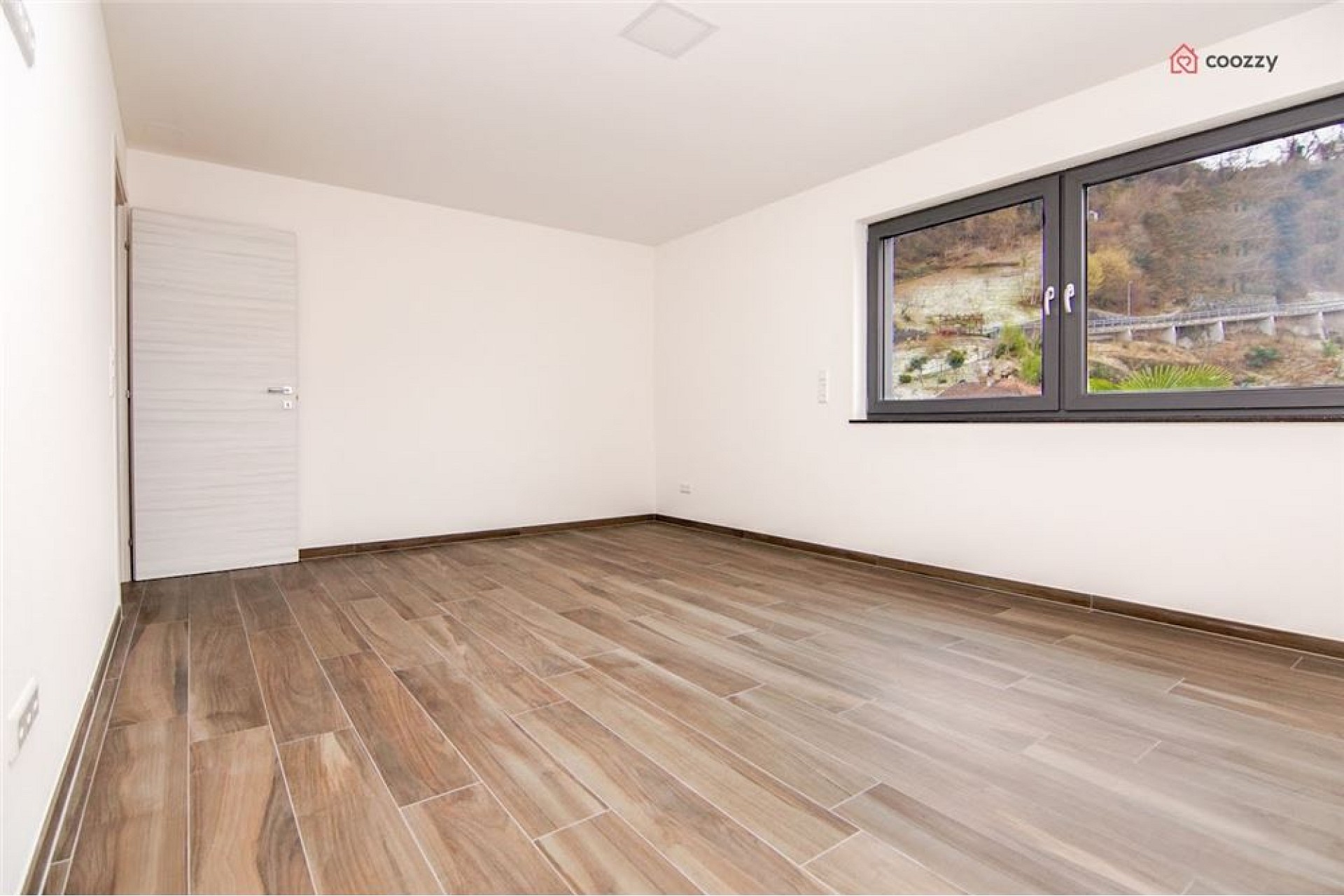 Leerer heller Raum mit Holzoptik-Boden und großem Fenster