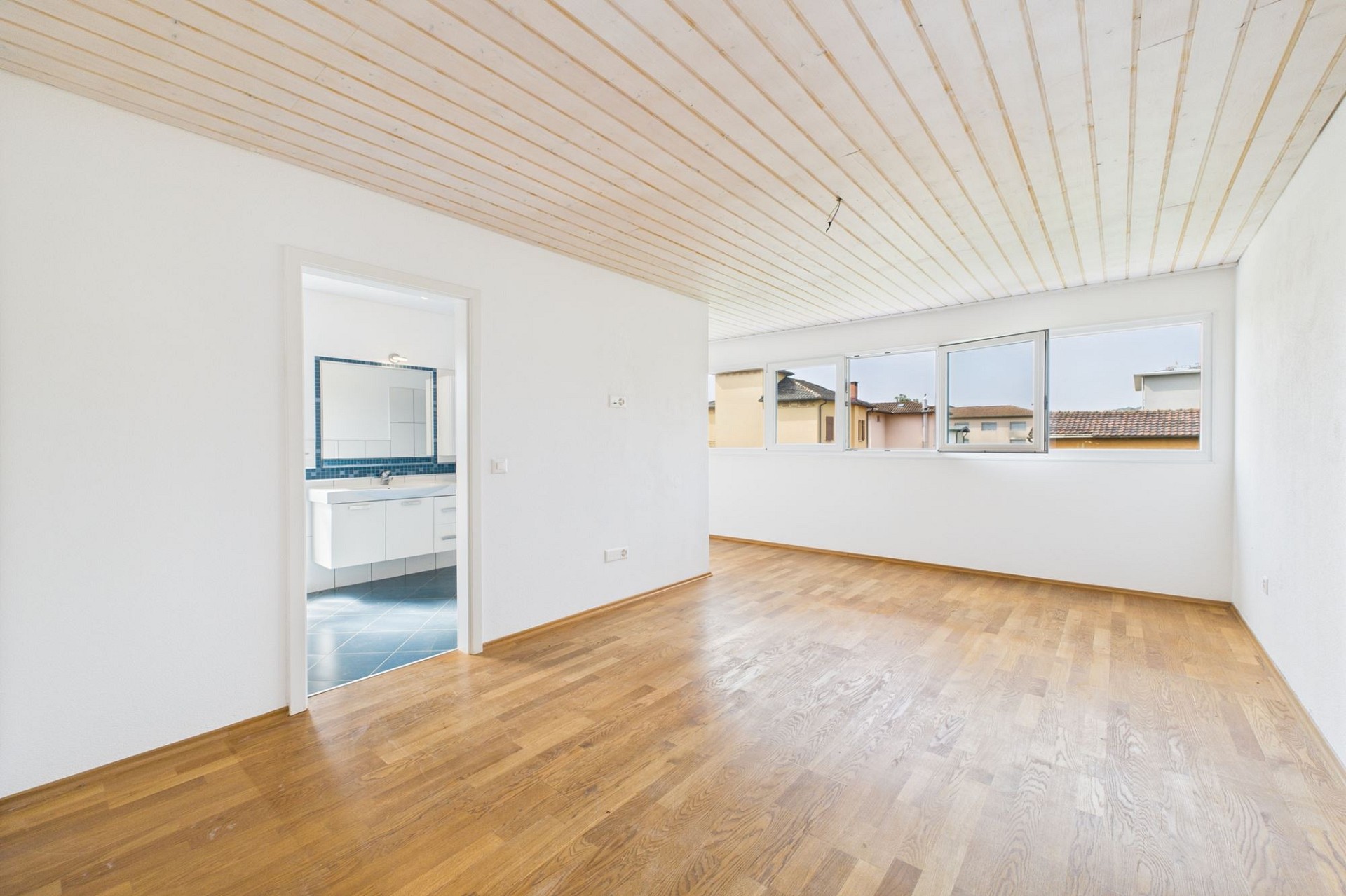 Heller leerer Wohnraum mit Blick ins angrenzende Badezimmer und auf die Fensterfront.
