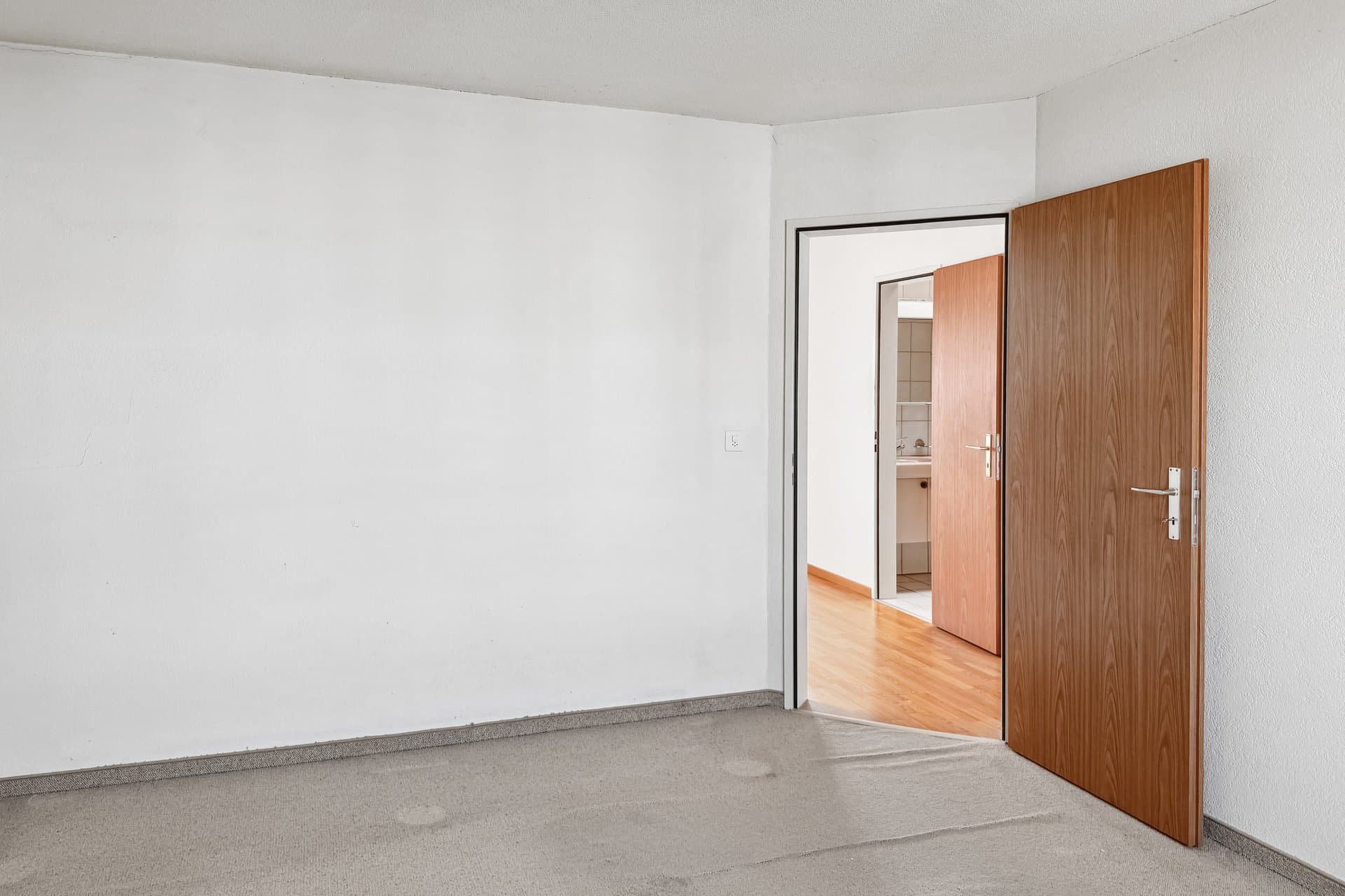 Ein schlichtes, teilmöbliertes Zimmer mit Blick in einen angrenzenden Flur oder Sanitärbereich.