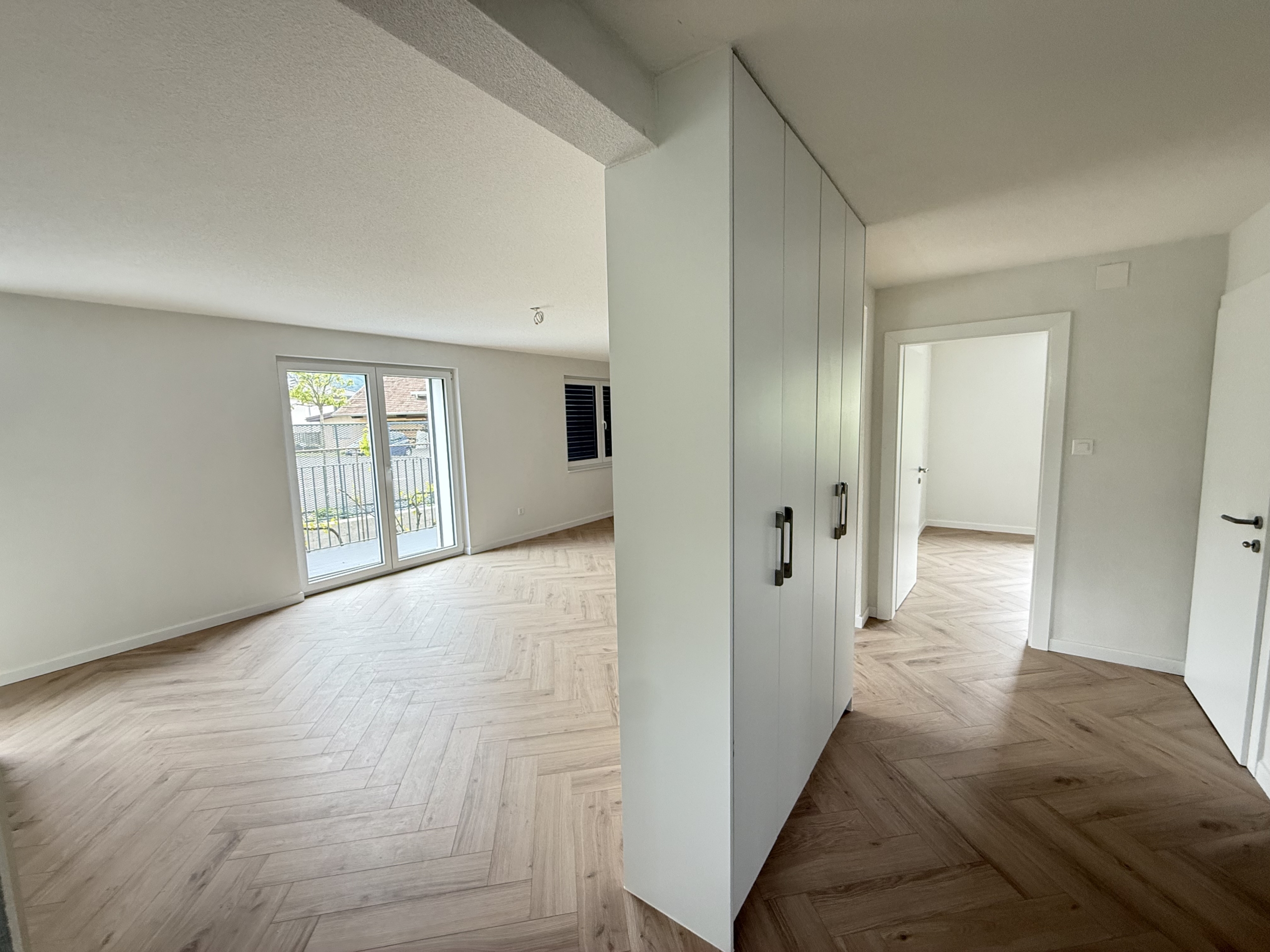 Helles, leeres Zimmer mit Fischgrätparkett und bodentiefer Tür zum Balkon