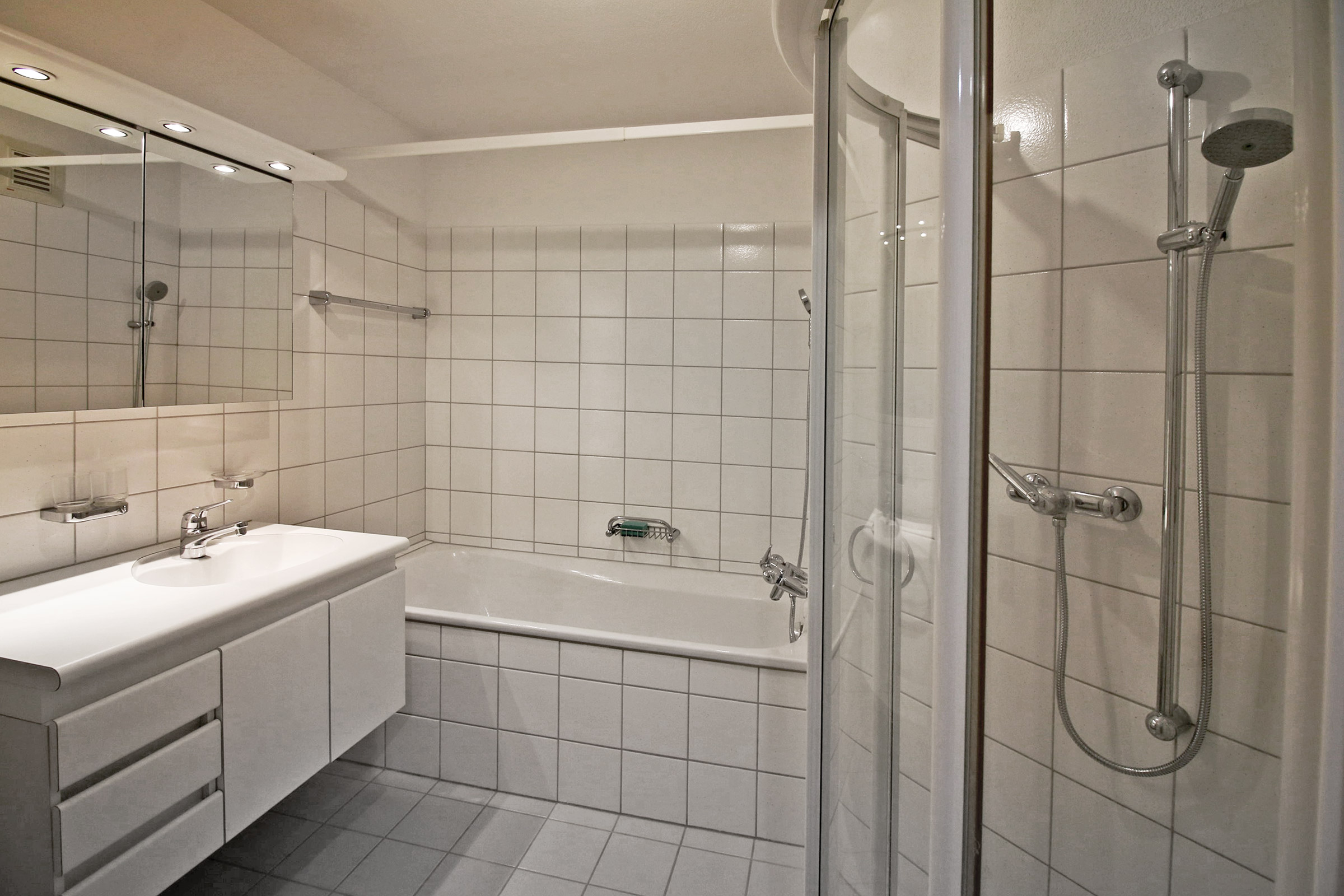 Helles, weiß gefliestes Badezimmer mit Badewanne und Duschbereich