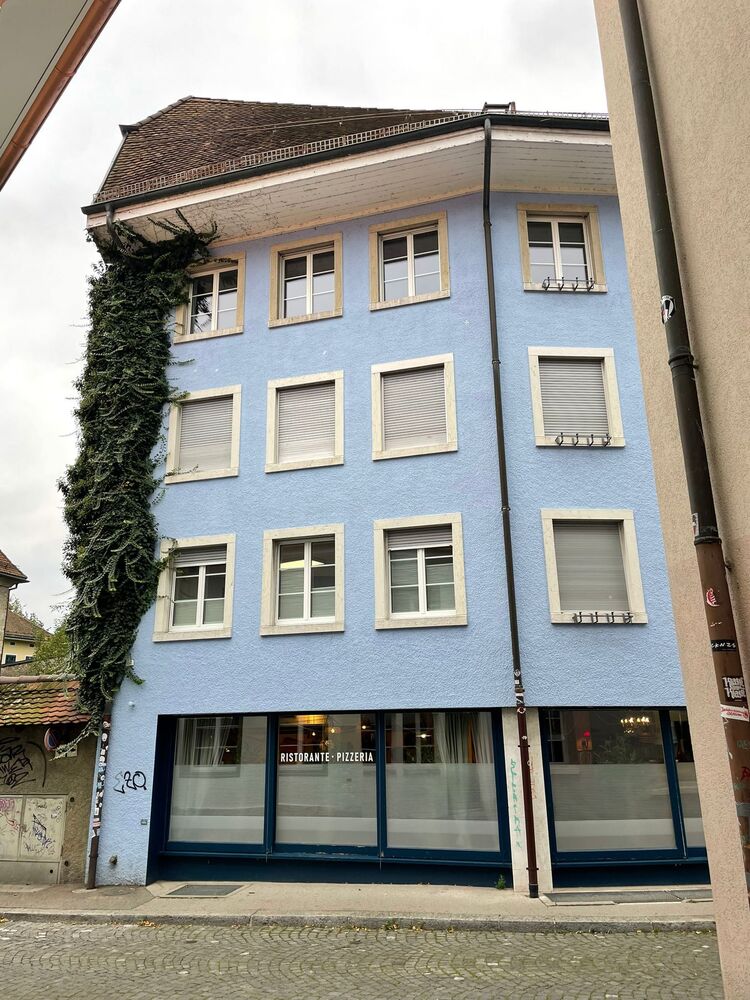 Straßenansicht eines blau verputzten Mehrfamilien- oder Geschäftshauses mit Ladenlokal im Erdgeschoss