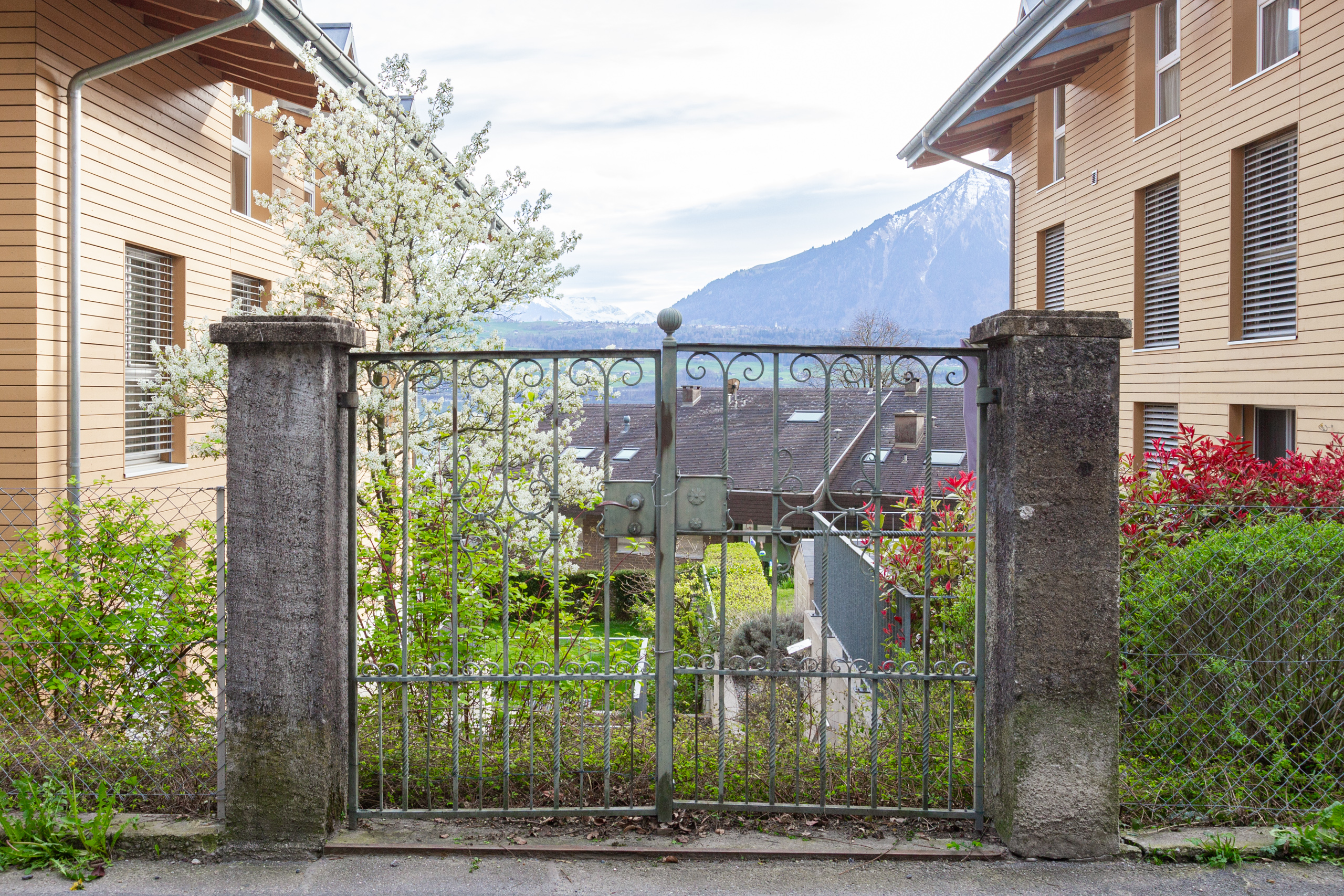 Blick durch ein Gartentor auf Wohngebäude und Bergkulisse