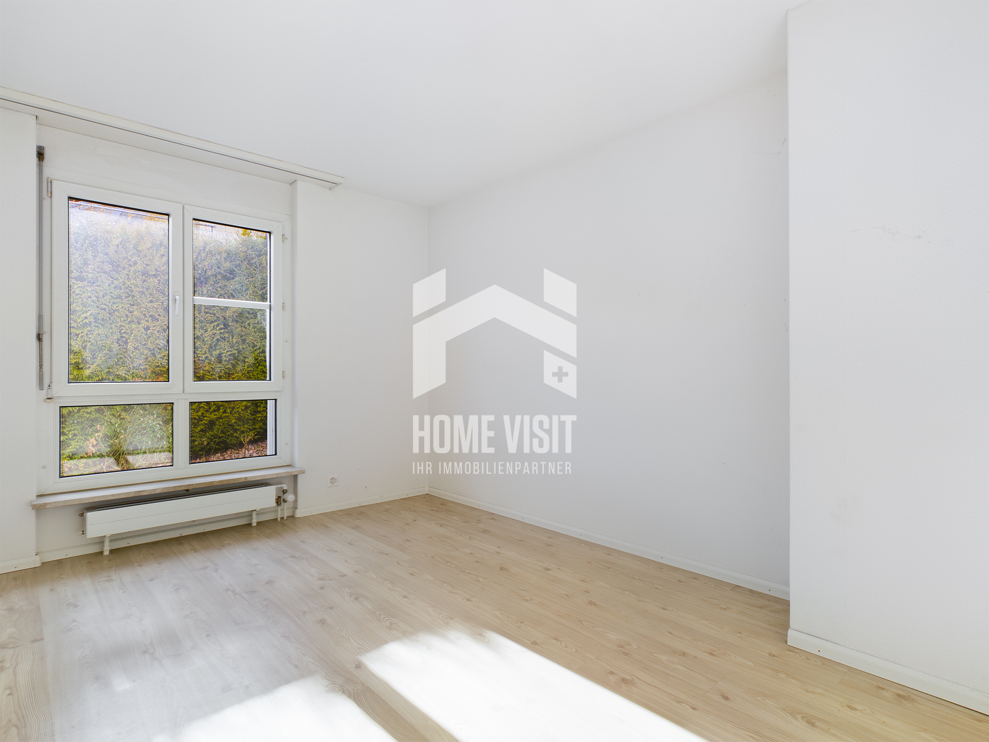 Helles, leeres Zimmer mit großem Fenster und hellem Holzboden
