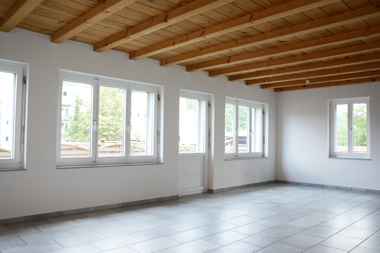 Leerer Wohnraum mit sichtbaren Holzdeckenbalken und mehreren Fenstern