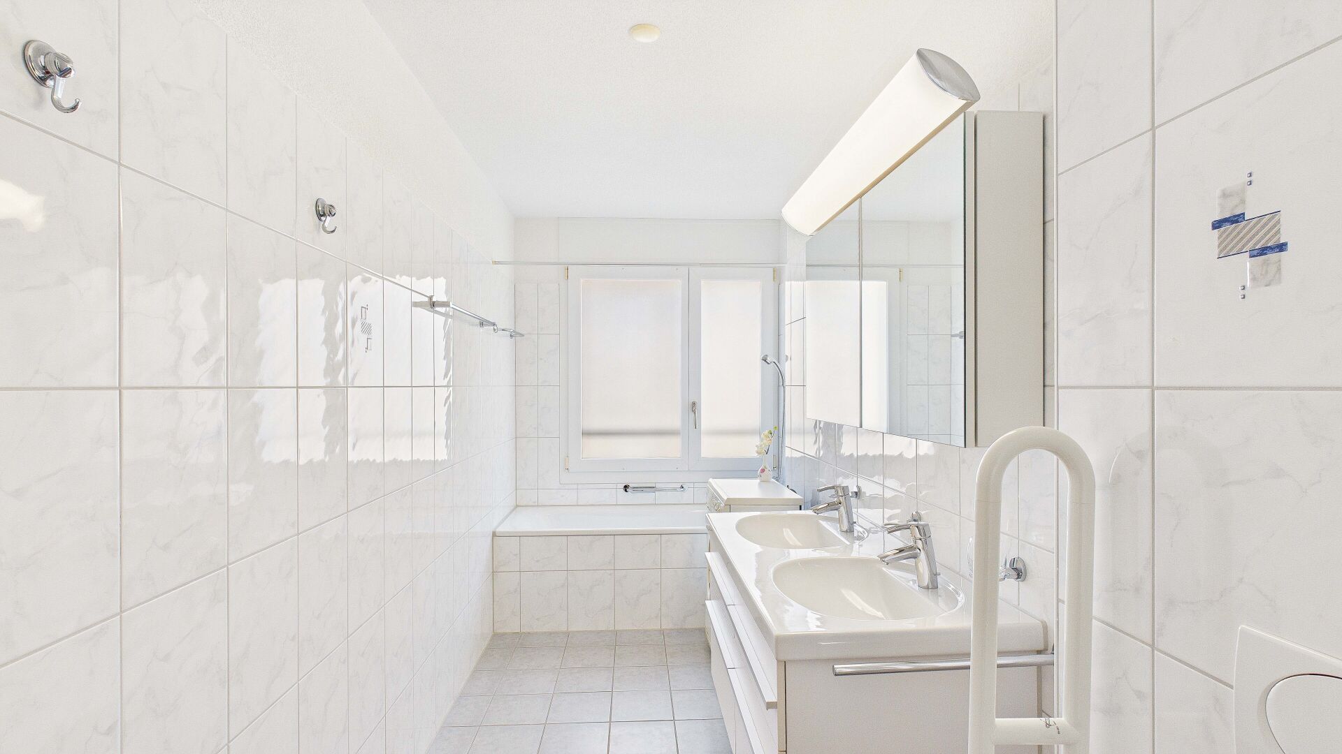 Helles weiß gefliestes Badezimmer mit Doppelwaschbecken und Badewanne