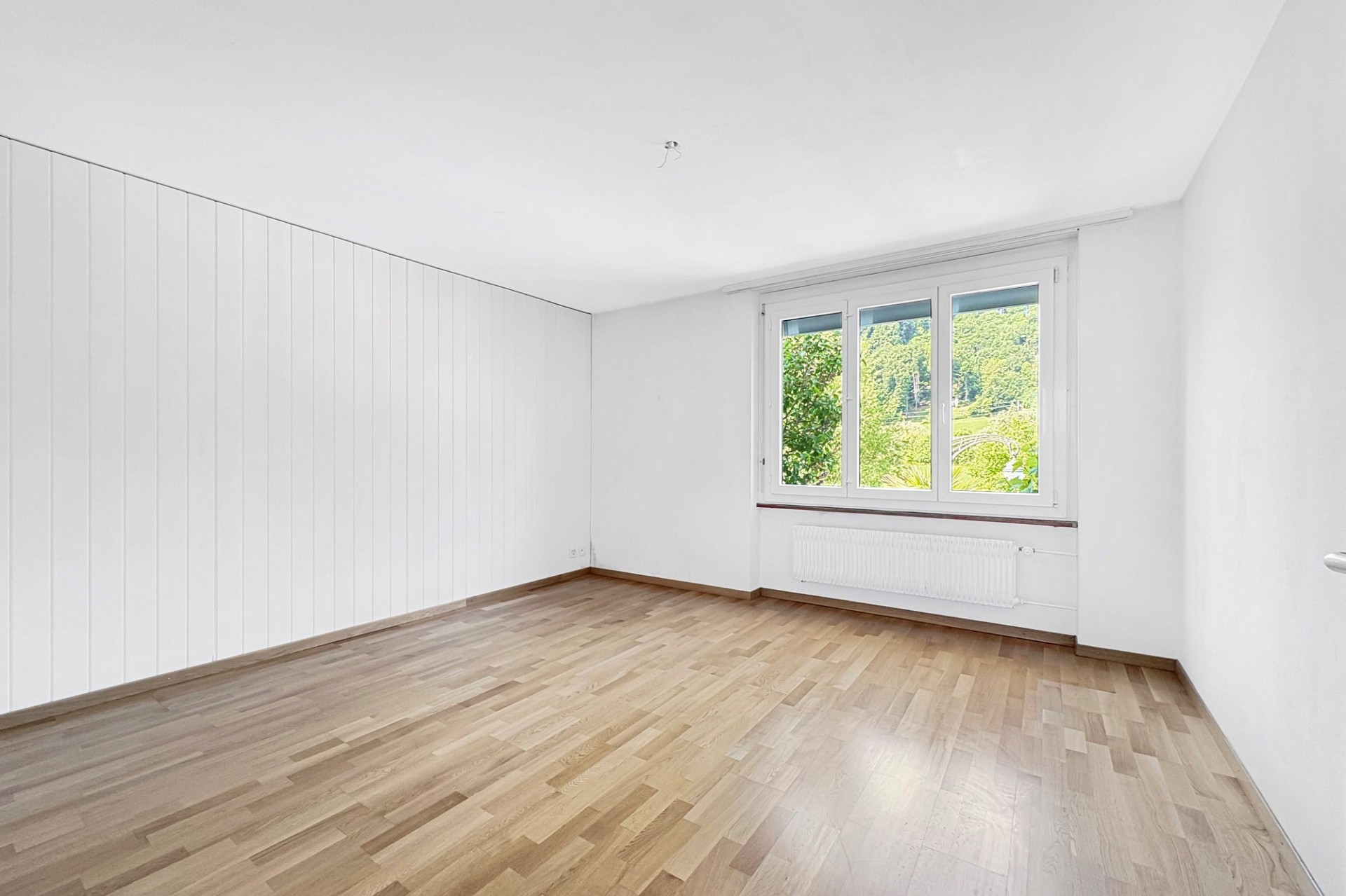Helles, leeres Zimmer mit Holzoptikboden und Fensterblick ins Grüne