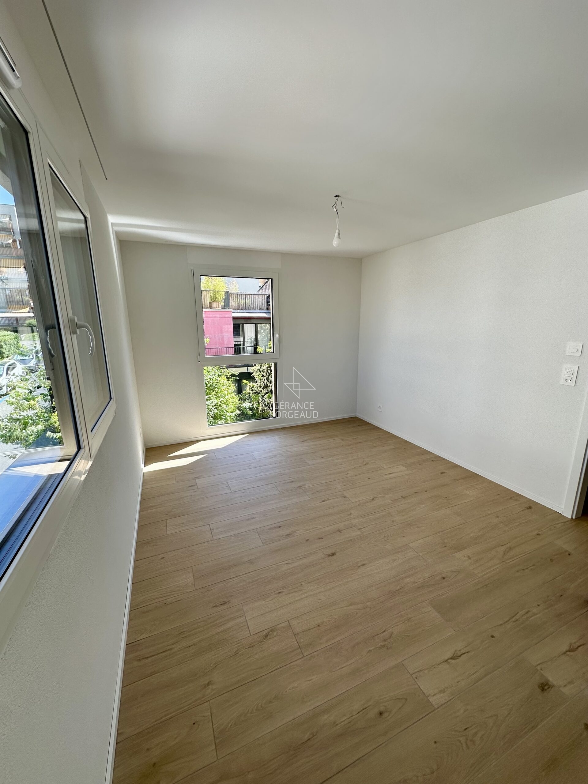 Helles, leeres Zimmer mit Holzoptikboden und zwei Fenstern