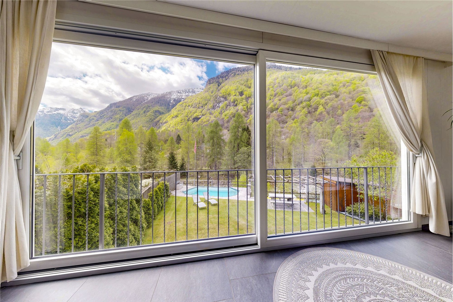 Blick aus einem Wohnraum auf Balkon, Pool und Berglandschaft