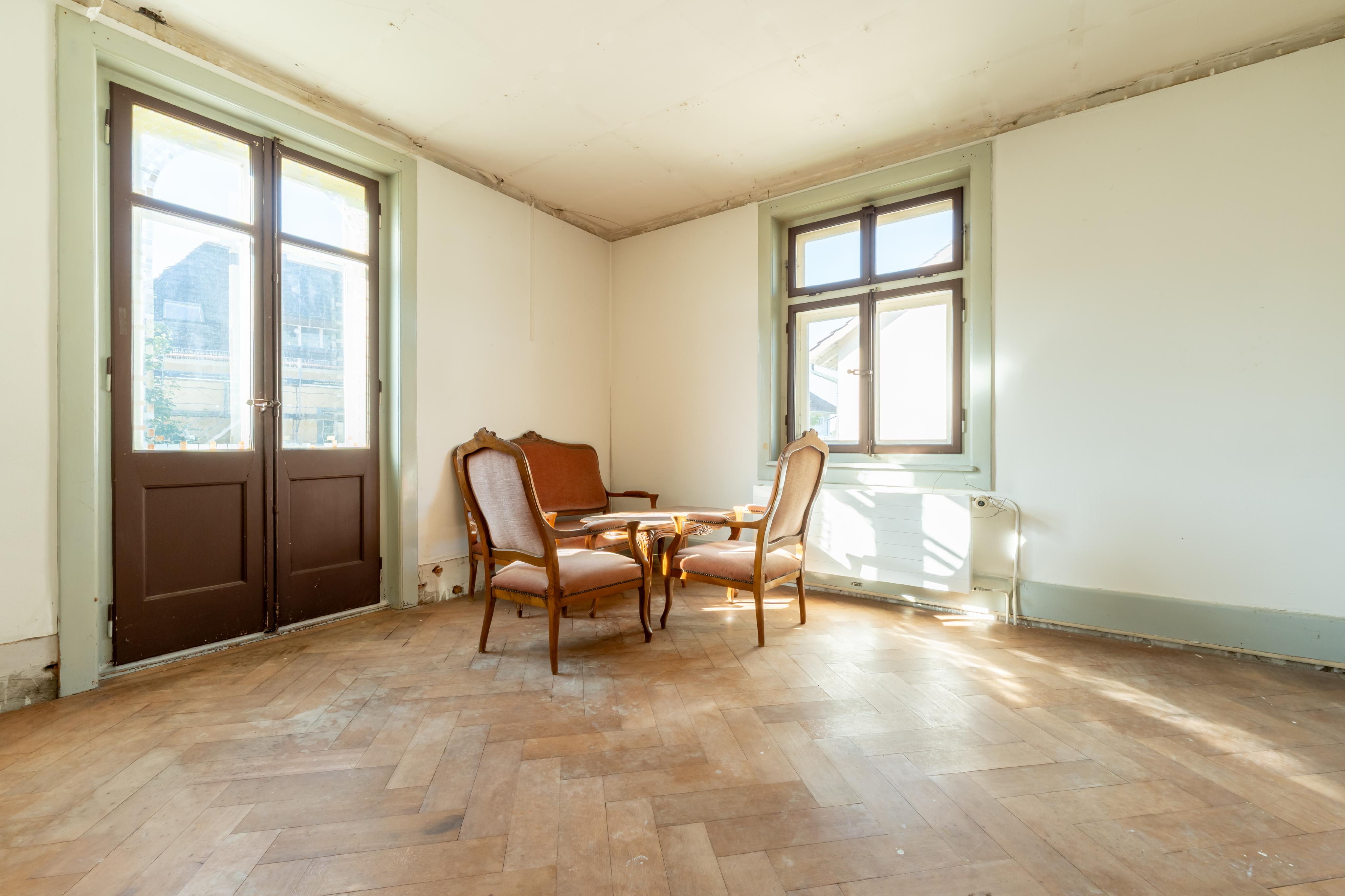 Heller, leerer Altbauraum mit zwei Fenstern und Holzstühlen