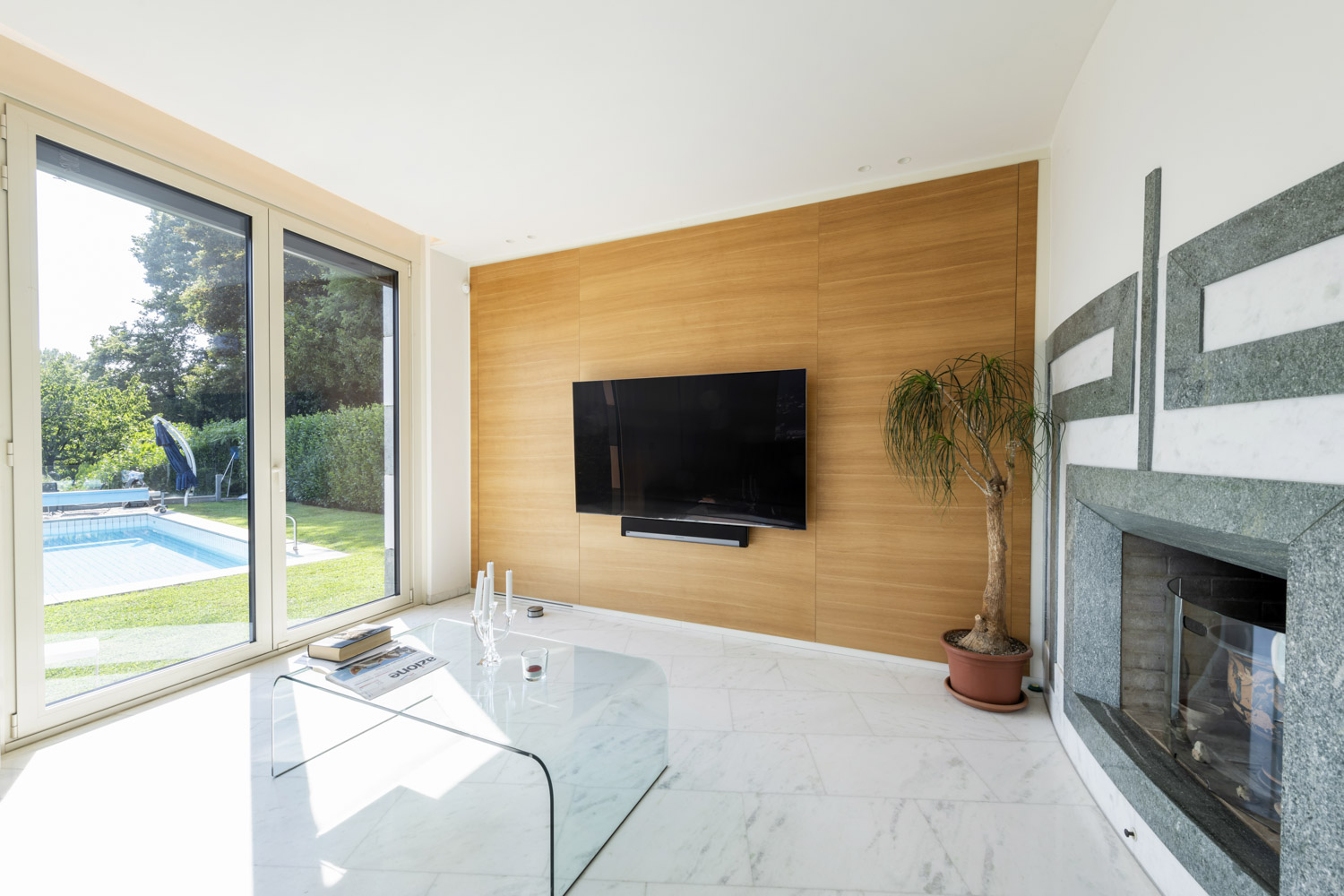 Heller Wohnraum mit Kamin, großer Glasfront und Blick auf den Garten mit Pool