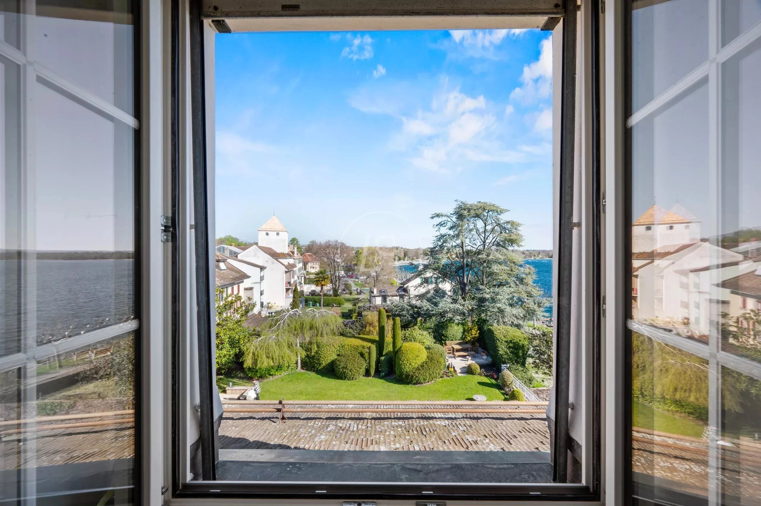 Blick durch ein geöffnetes Fenster auf Wasser, Garten und Uferbebauung