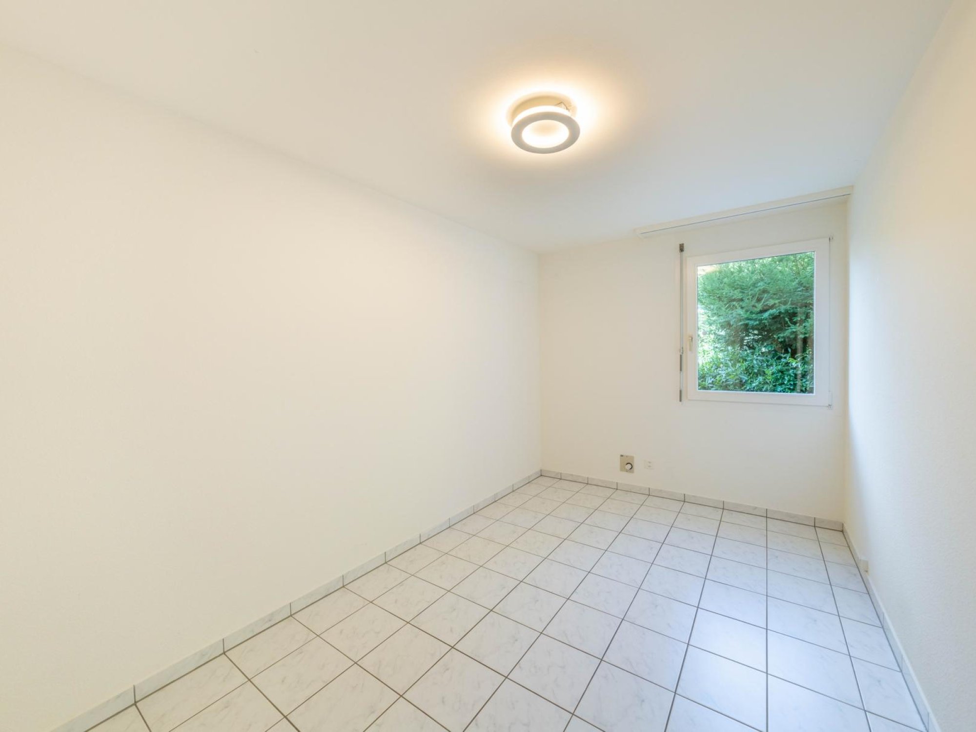 Heller leerer Raum mit Fliesenboden und Fensterblick ins Grüne