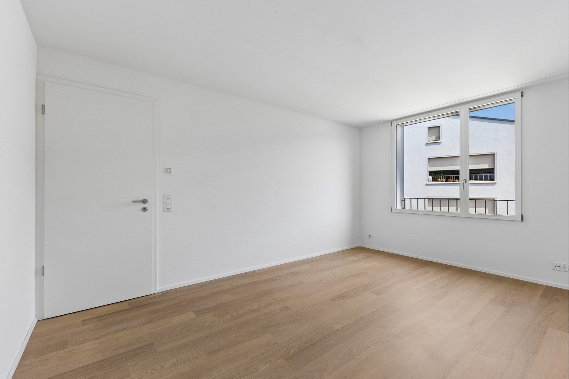 Helles, leeres Zimmer mit Holzoptikboden und Fensterblick in den Innenhof.